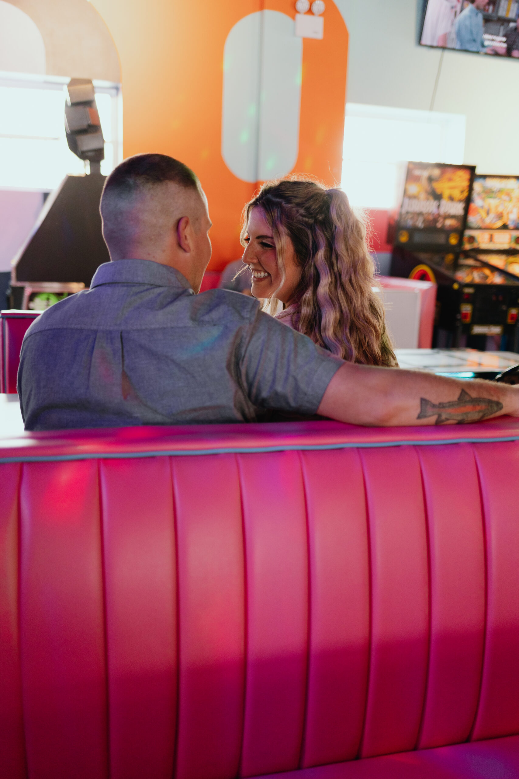 A couple shares a joyful moment together at a vibrant arcade bar, with the woman laughing as she looks at the man, seated in a classic red booth with a colorful, playful background setting the scene.