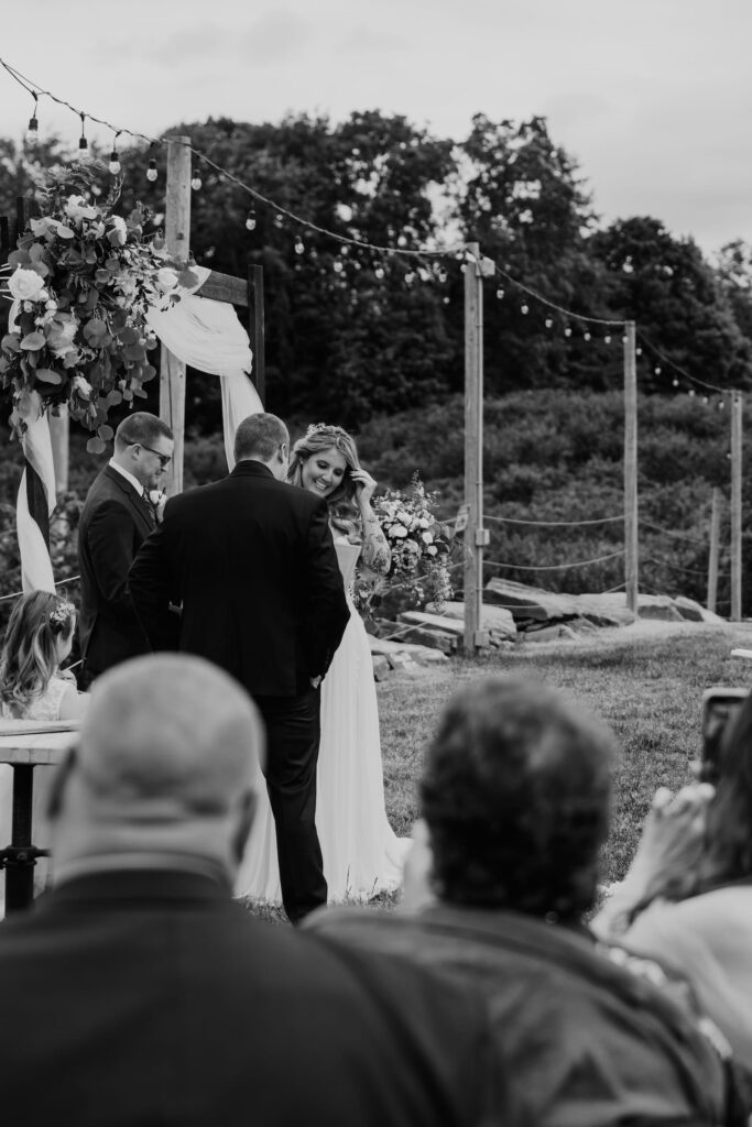 Bride and groom stand at the altar with a rustic wooden arch and string lights overhead, exchanging smiles during their wedding ceremony.