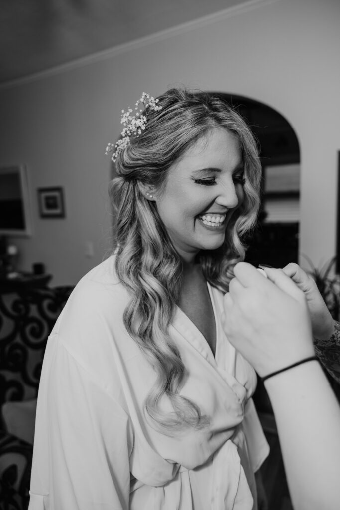 A joyful bride in a white robe with wavy hair and a delicate floral hairpiece smiles brightly, capturing a candid moment of happiness before the wedding ceremony.