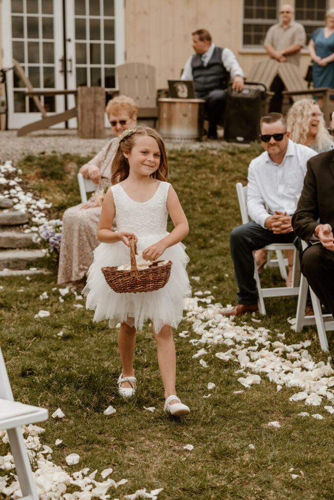 A flower girl in a white dress with a lacy top and tulle skirt walks down the aisle scattering petals from a wicker basket.