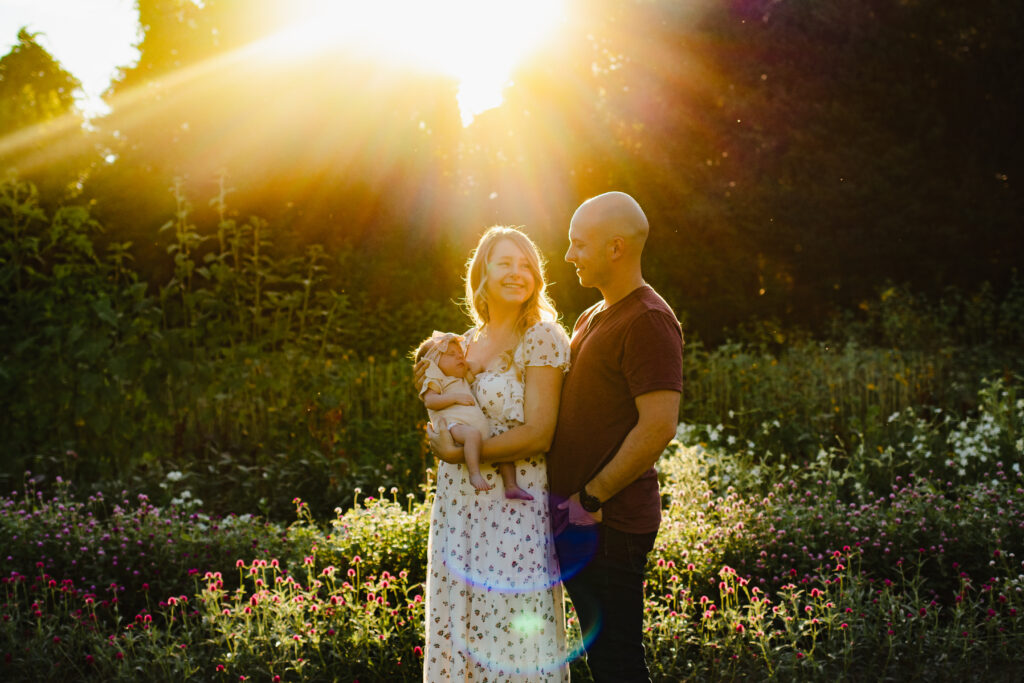 In a golden field at sunset, a mother lovingly cradles her newborn, while the father looks on adoringly, their silhouettes framed against the radiant sunlight filtering through the trees.