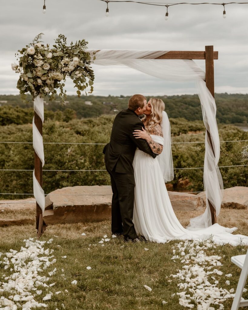 A newlywed couple shares a romantic kiss under a floral-decorated wedding arch, with sheer drapery blowing gently and a scenic green landscape in the background.