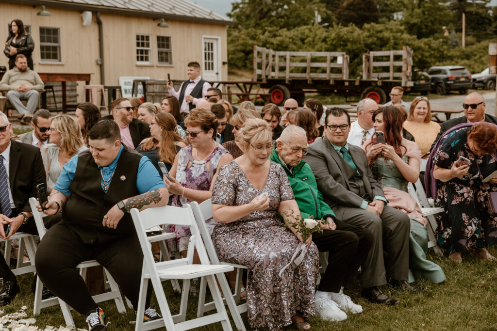 Guests seated in white chairs at an outdoor wedding ceremony, attentively waiting for the event to start, with a rustic barn and vintage wagon in the background.