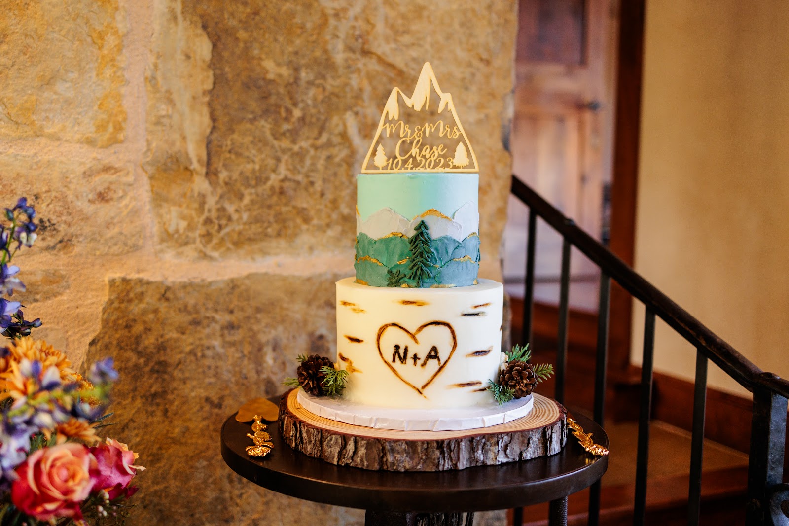 Two-tiered adventurous wedding cake with mountain and sky icing art on the upper tier, a heart with initials 'N+A' on the lower tier, and a personalized 'Mr & Mrs Chase' topper, displayed on a wood slice stand amidst flowers and pine cones.