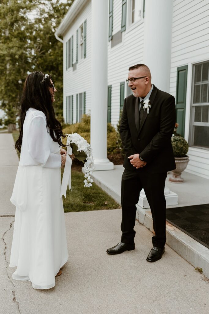 A bride in a white dress with a floral headpiece laughs with the groom in a black suit, both standing in front of a white colonial house with green shutters.