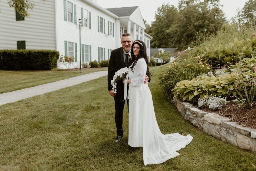 Bride in a flowing white dress and groom in a classic black suit standing together on a lush green lawn, with a charming white colonial house and vibrant garden in the background.