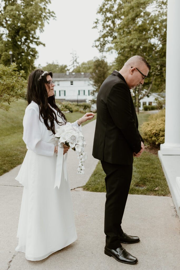 A bride in a white dress with a long-sleeved cape and holding a bouquet stands beside a groom in a black suit, both looking away from the camera, with a colonial-style house and greenery in the background at Curry Estate.