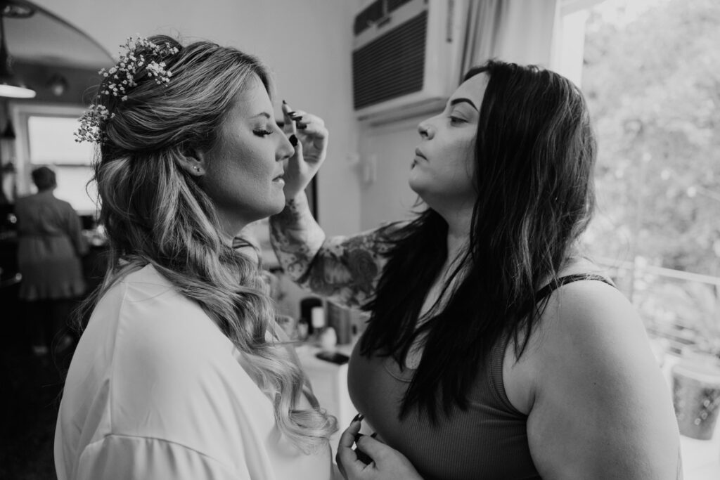 Bride receiving final makeup touches before the wedding, with her maid of honor assisting, sharing a moment of anticipation and joy.