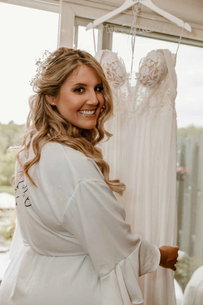 Radiant bride in her personalized robe smiling over her shoulder, with her wedding dress hanging in the background, ready for her special day at Locust Grove.
