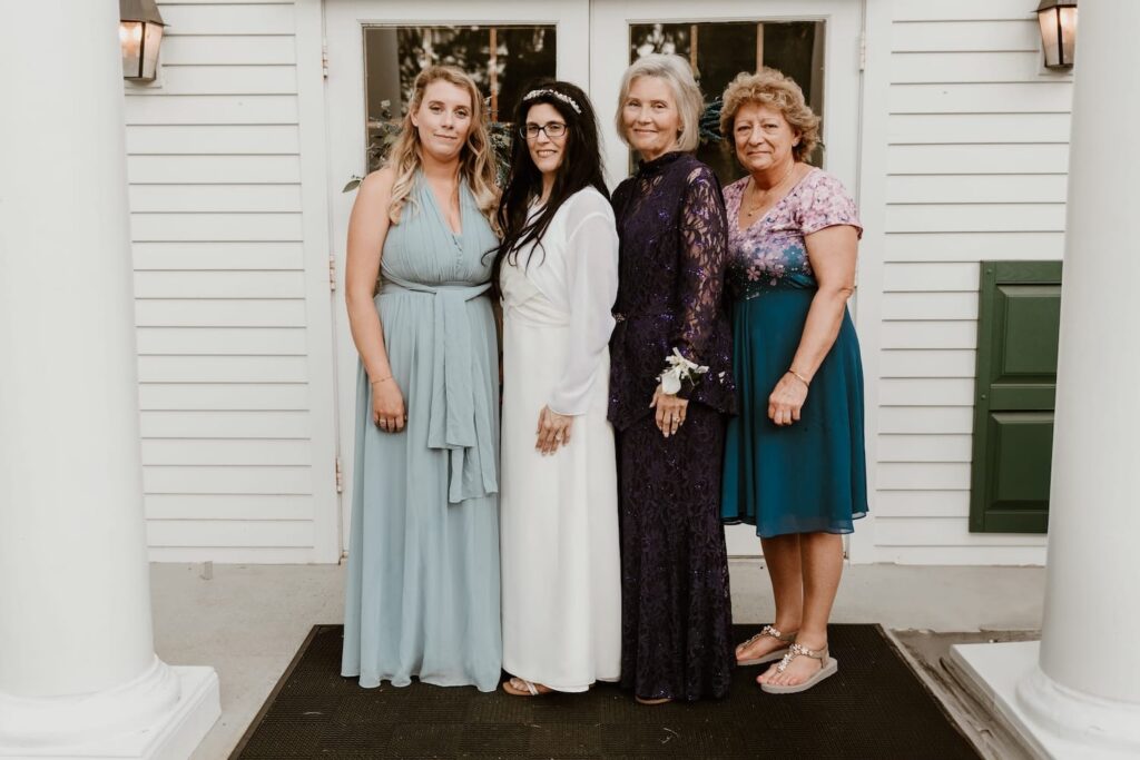 A bride in a white dress and crystal headpiece stands with three women wearing elegant dresses in shades of blue and purple, posing together on a porch.