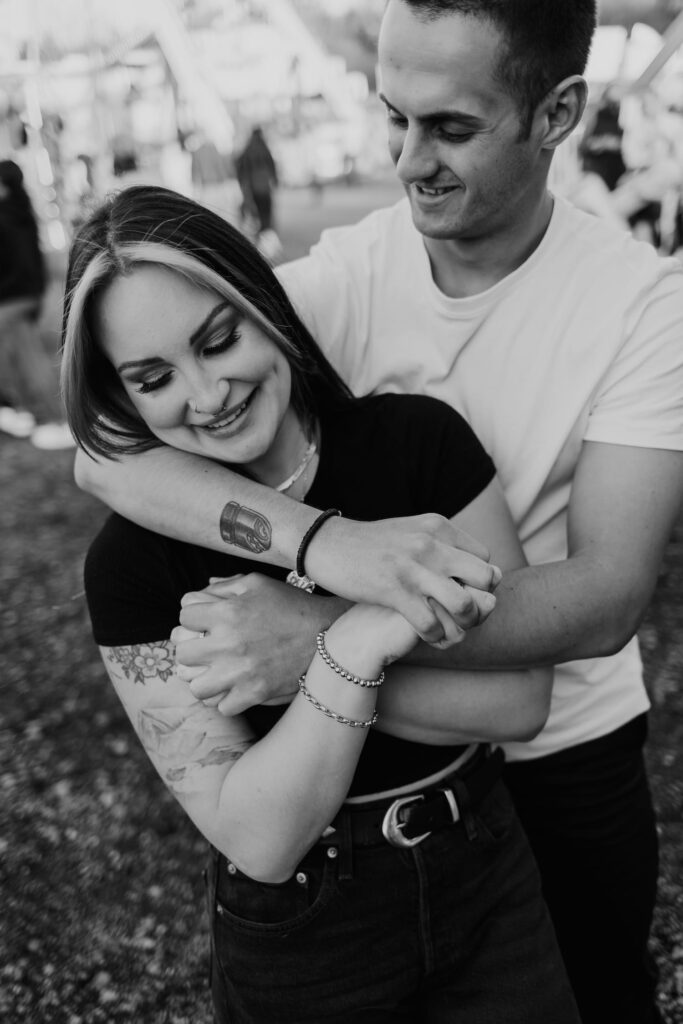 A monochrome image captures a tender moment as a man wraps his arms around a woman, both smiling, at the Dutchess County Stadium Carnival.
