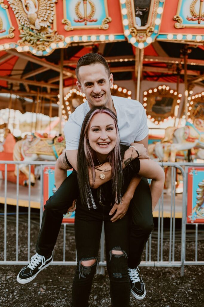 A couple shares a playful moment at the Dutchess County Stadium Carnival, with colorful carousel lights twinkling in the background.