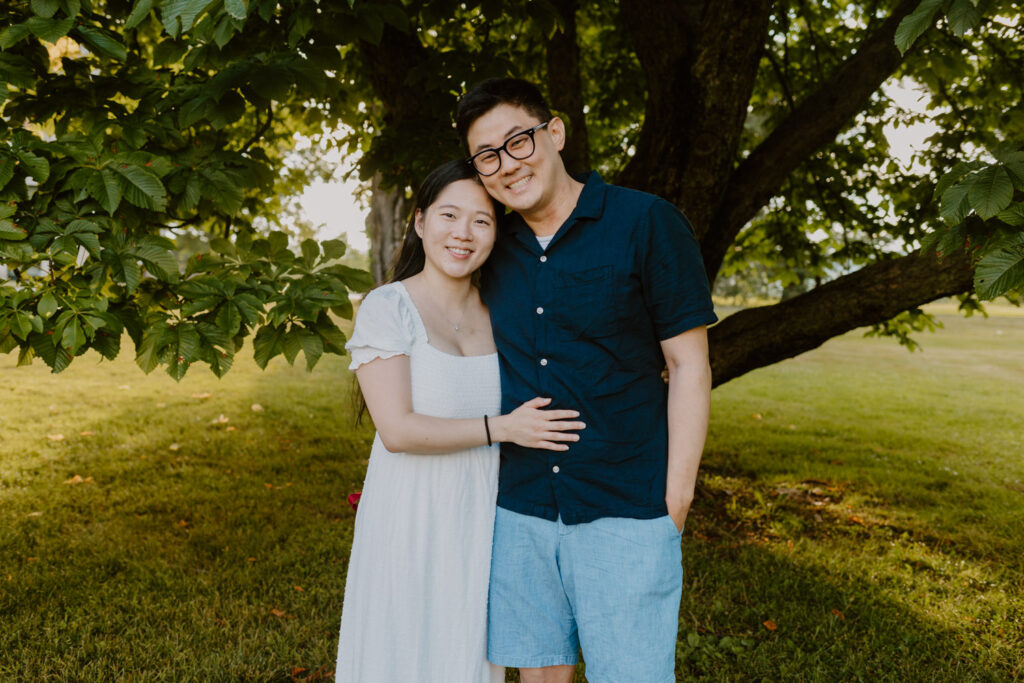 A couple stands closely, embracing under the shade of a lush tree, with sunlight filtering through the leaves. The woman, dressed in a white summer dress, leans against the man who is smiling broadly in a navy blue button-up shirt and glasses.