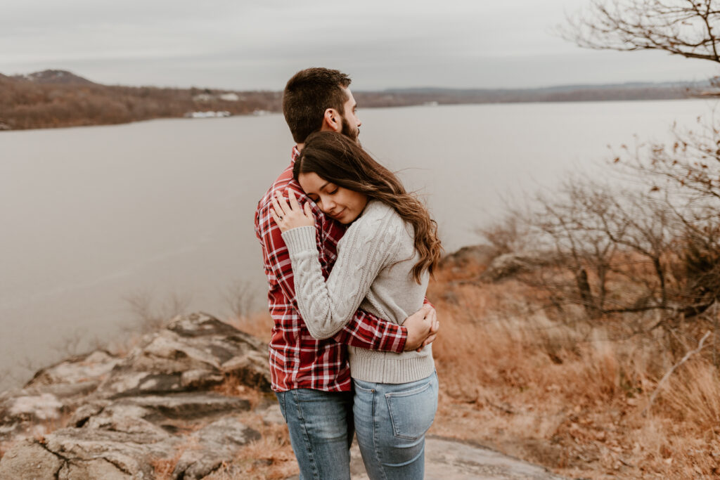 A couple embraces lovingly atop Breakneck Ridge, overlooking the Hudson River, sharing a moment of connection amidst the tranquil, scenic landscape.