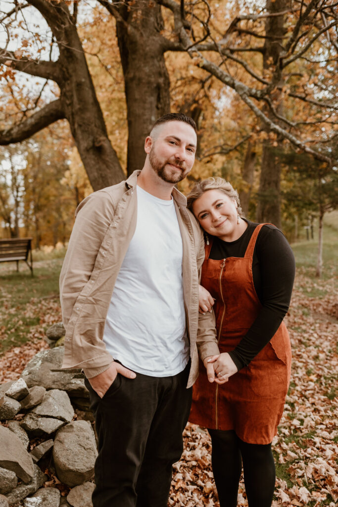A couple stands close together, smiling contentedly, against a backdrop of autumn leaves and a majestic tree in a park.