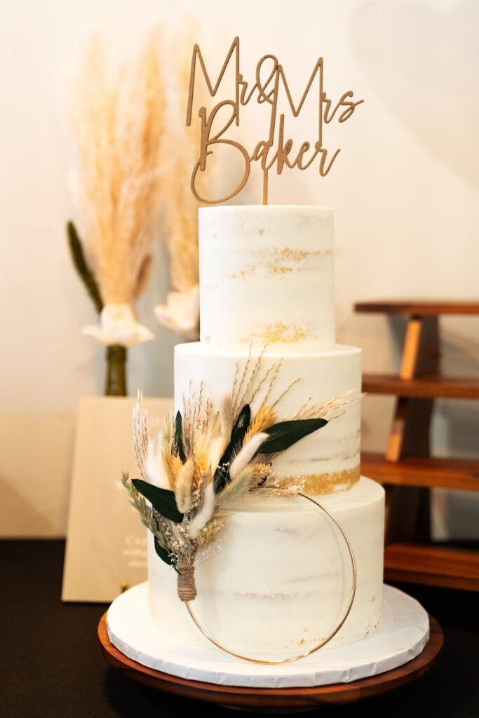 Elegant ivory three-tier wedding cake with gold marbling and a natural dried flower arrangement, featuring a custom 'Mr & Mrs Baker' topper, against a soft backdrop.