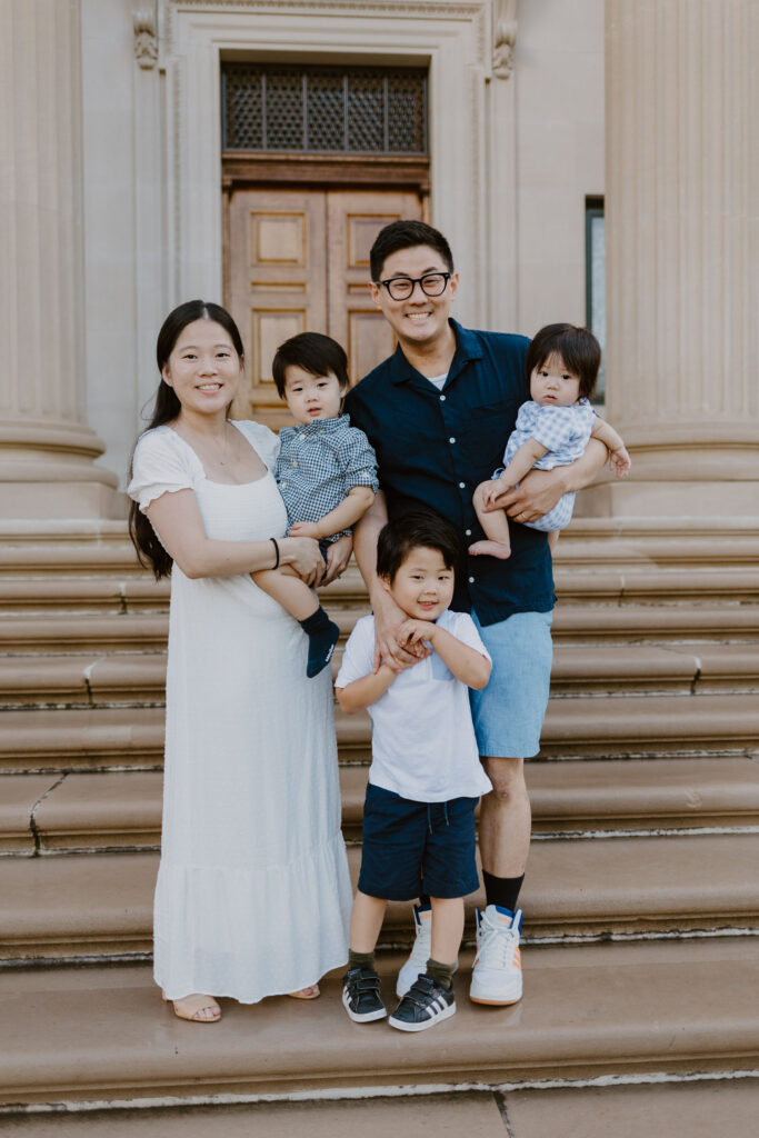 A happy family of five poses on the steps of a stately building. The mother, in a white dress, holds their youngest in a plaid shirt, while the father in a navy blue shirt holds their other toddler. Their eldest child, wearing a white shirt and navy shorts, stands in front, making a playful pose.