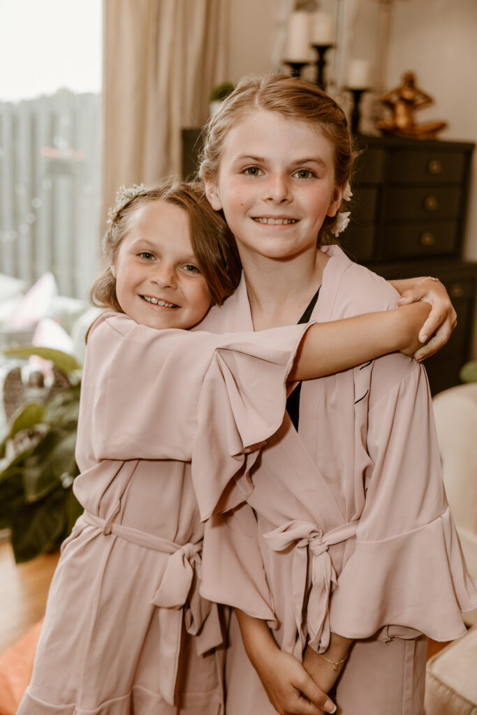 Two young flower girls in matching blush dresses sharing a warm embrace, filled with excitement for the wedding day.