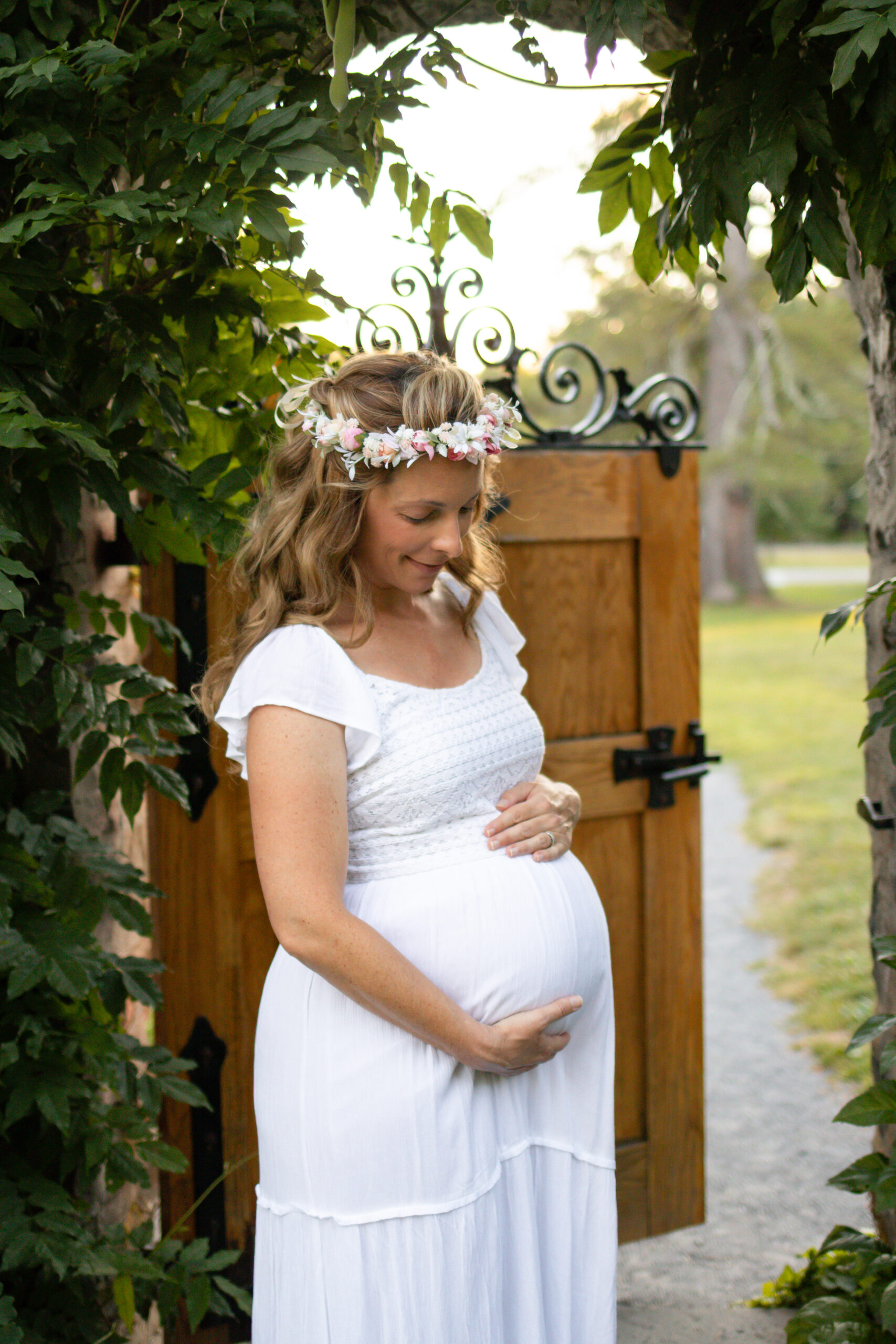 A serene pregnant woman in a white dress and floral headband stands gently cradling her belly under a leafy archway, with soft sunlight filtering through the foliage.