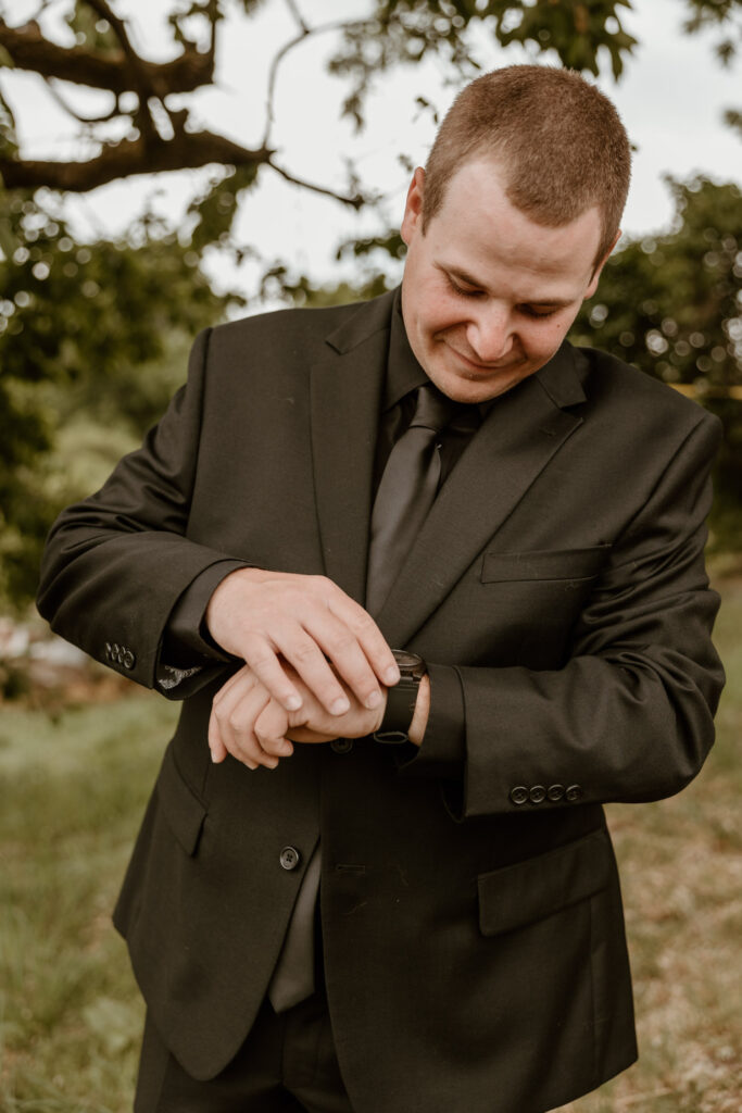 A groom in a black suit adjusts his watch outdoors with a serene expression, surrounded by natural greenery.