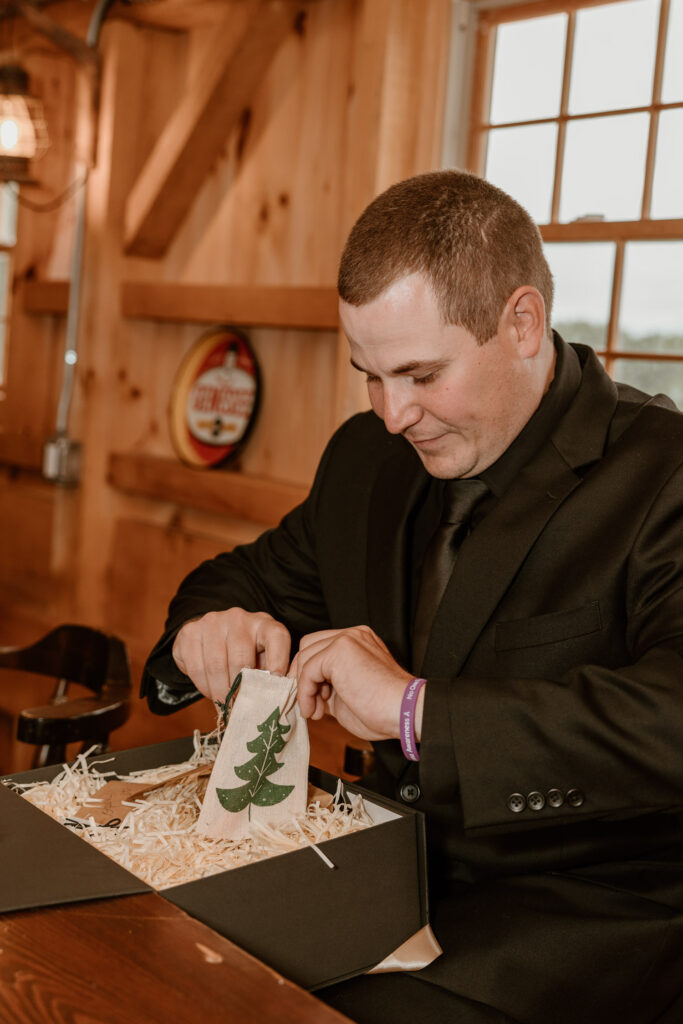 A groom in a classic black suit intently opens a small burlap gift bag with a green tree print, set against a rustic Locust Grove Brewery wooden interior.