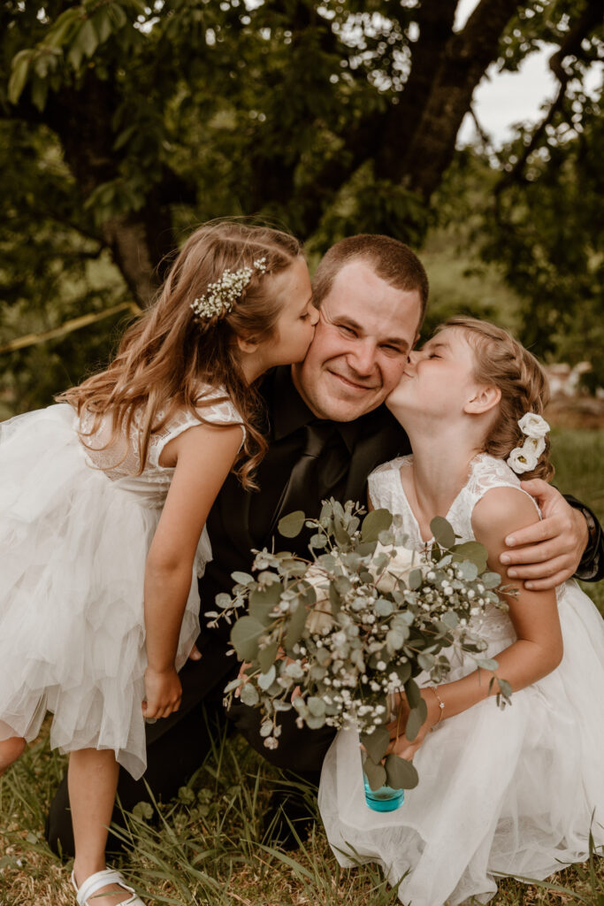 Joyful groom kneeling and getting kisses on the cheek from two young flower girls, capturing a sweet moment at a wedding.