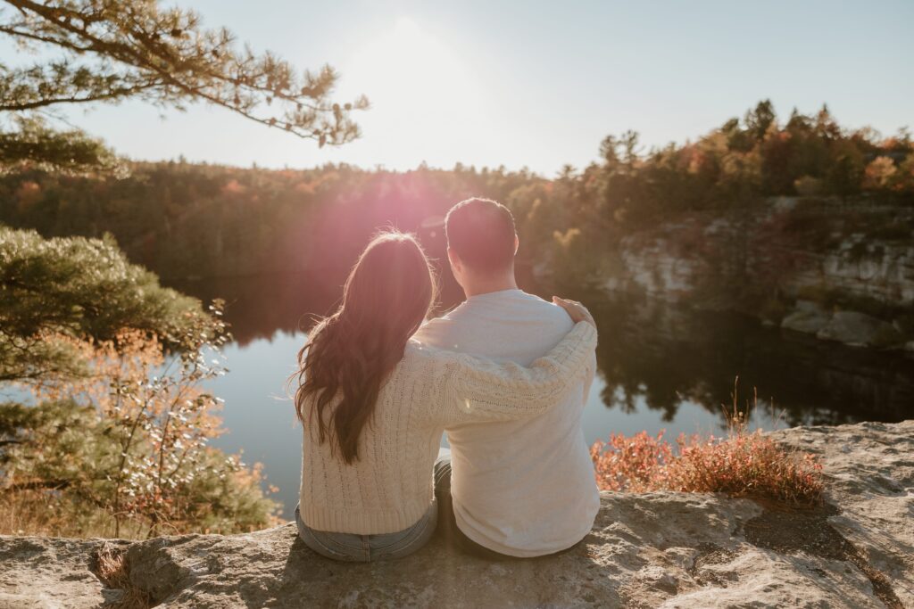 Couple sitting on a cliff at Lake Minnewaska, embracing as the sun sets over the scenic autumn landscape.