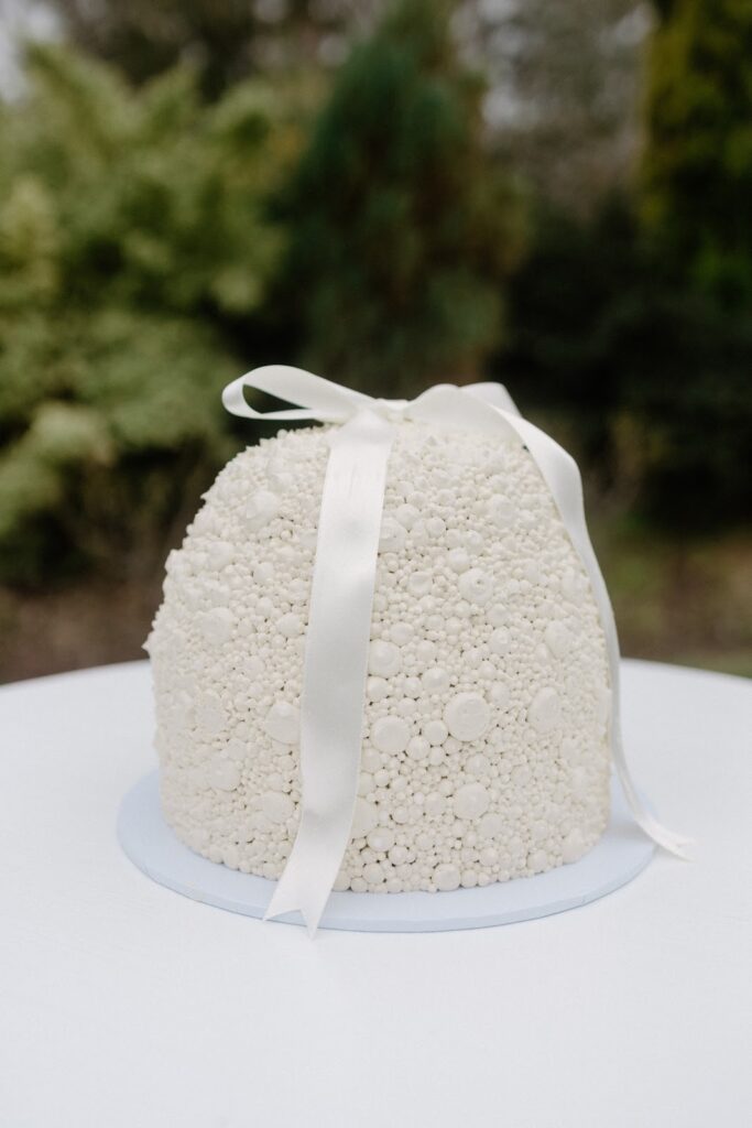 Minimalist ivory dome-shaped wedding cake decorated with sugar pearls and finished with a satin ribbon, showcasing an elegant and modern design.