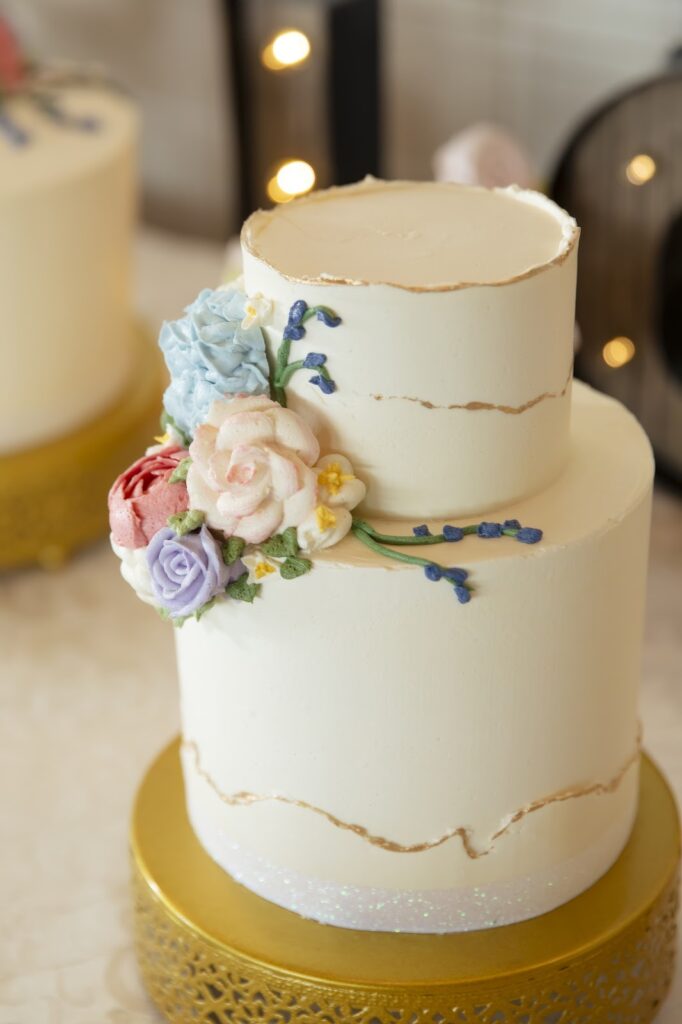 Elegant two-tier wedding cake with gold trim and a cascade of delicate pastel flowers, presented on a golden pedestal stand, perfect for a romantic wedding reception.
