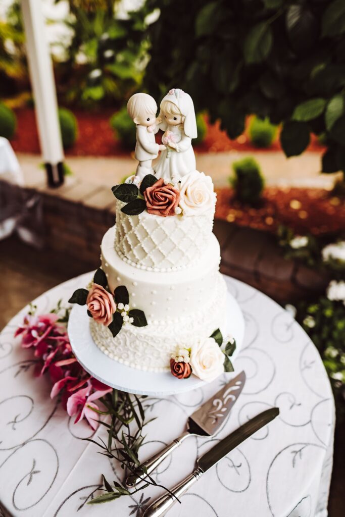 An exquisite three-tier wedding cake with lace detailing and pastel rose embellishments, crowned with a bride and groom figurine, set against a backdrop of lush foliage.