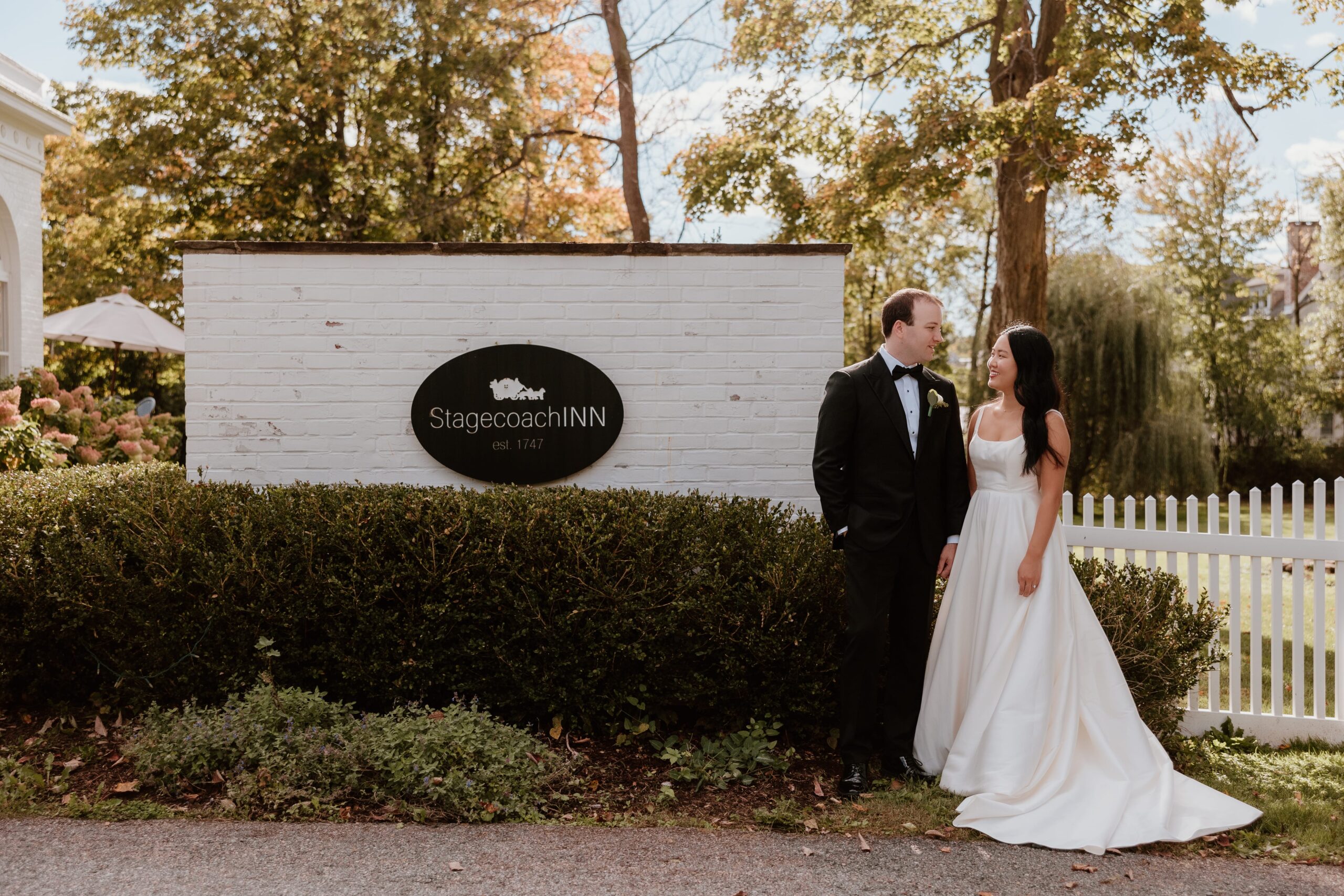 A bride and groom standing in front of the iconic Stagecoach Inn sign, located in Goshen, NY. The bride is wearing a classic white gown, and the groom is dressed in a sharp black tuxedo. They are smiling at each other against a backdrop of lush greenery, a white picket fence, and charming outdoor landscaping on a sunny day.