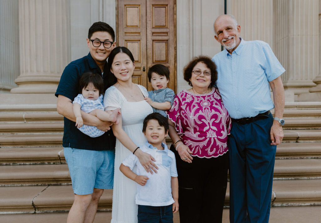 A multi-generational family poses together on the steps of Vanderbilt Mansion, with the parents holding their young children and flanked by their own parents, who are smiling proudly next to their grandchildren.