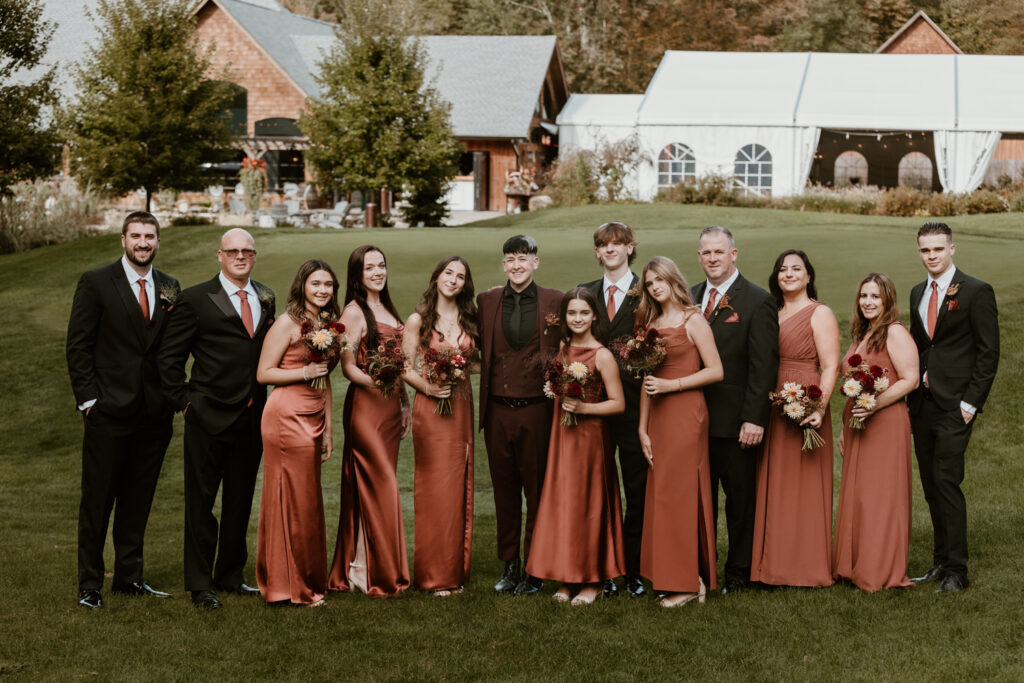 A wedding party in coordinated rust-colored attire posing on a lush green lawn with a rustic venue and white tent in the background at a hudson valley wedding venue.
