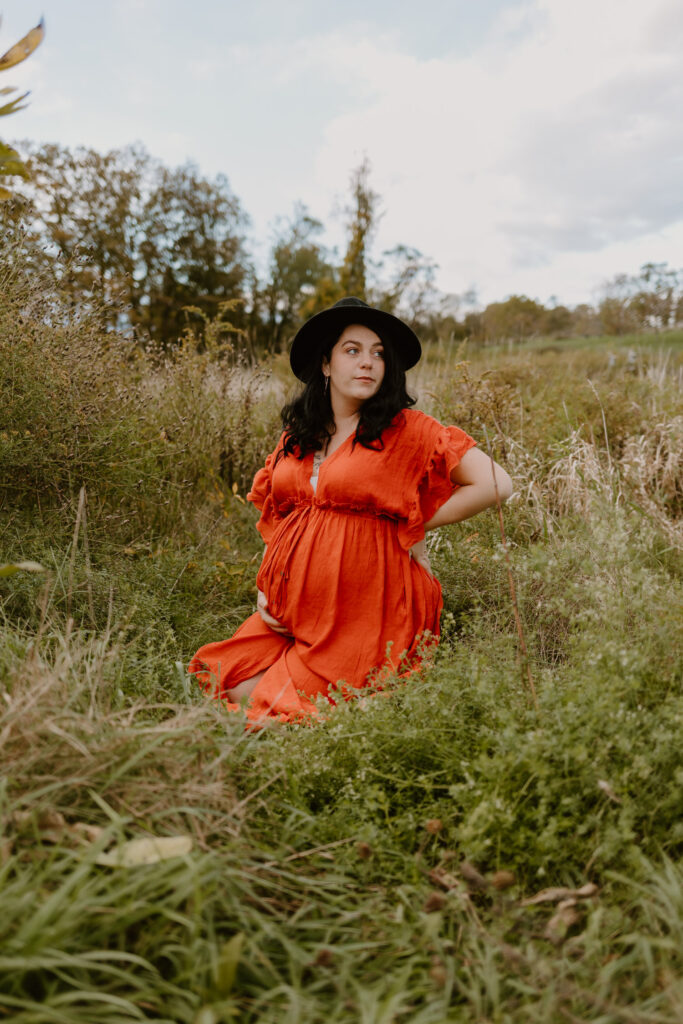 A woman in a vibrant orange dress and a black hat sits amidst tall grass at Drayton Grant Park at Burger Hill, gazing thoughtfully into the distance, embodying a sense of calm and connection with nature.