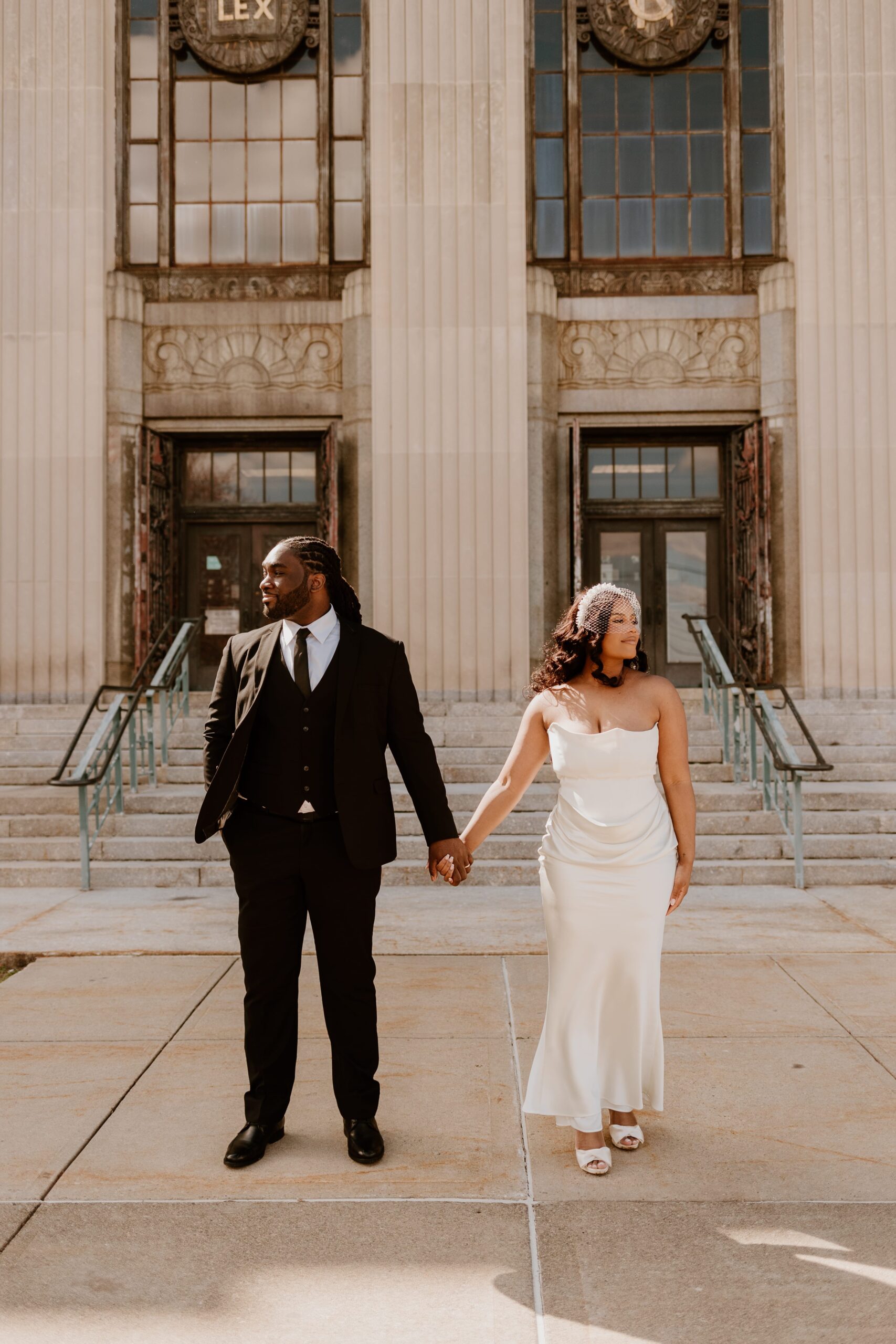 A newlywed couple holding hands in front of the grand entrance of Rockland County Courthouse, showcasing its historic architecture and the couple's elegant wedding attire.