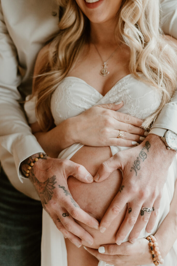 Close-up of a pregnant woman's belly with hands forming a heart shape, showcasing her lace-trimmed dress, necklace, and the intricate tattoos on the man's arms.
