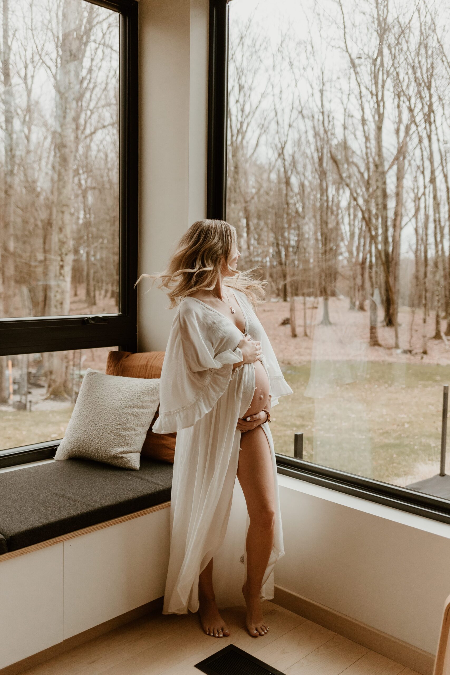A serene Hudson Valley maternity photographer captures a pregnant woman in a delicate white robe, standing by a large window overlooking a bare winter forest.
