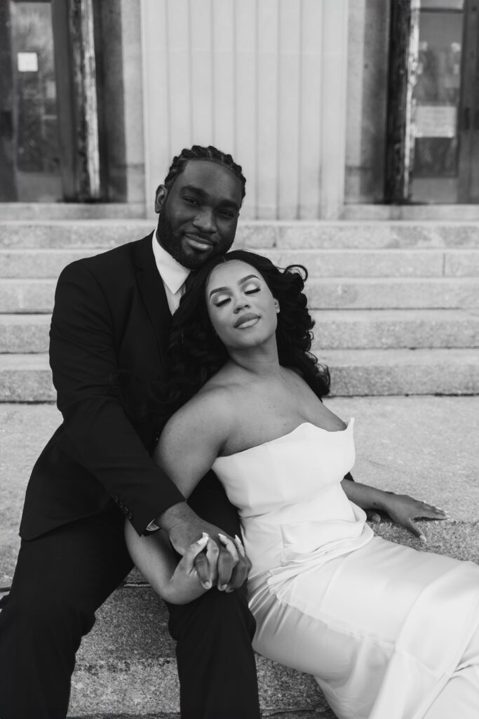 A tender black and white portrait of a groom seated on the courthouse steps with his bride reclining in his arms, capturing the romance of their courthouse wedding.