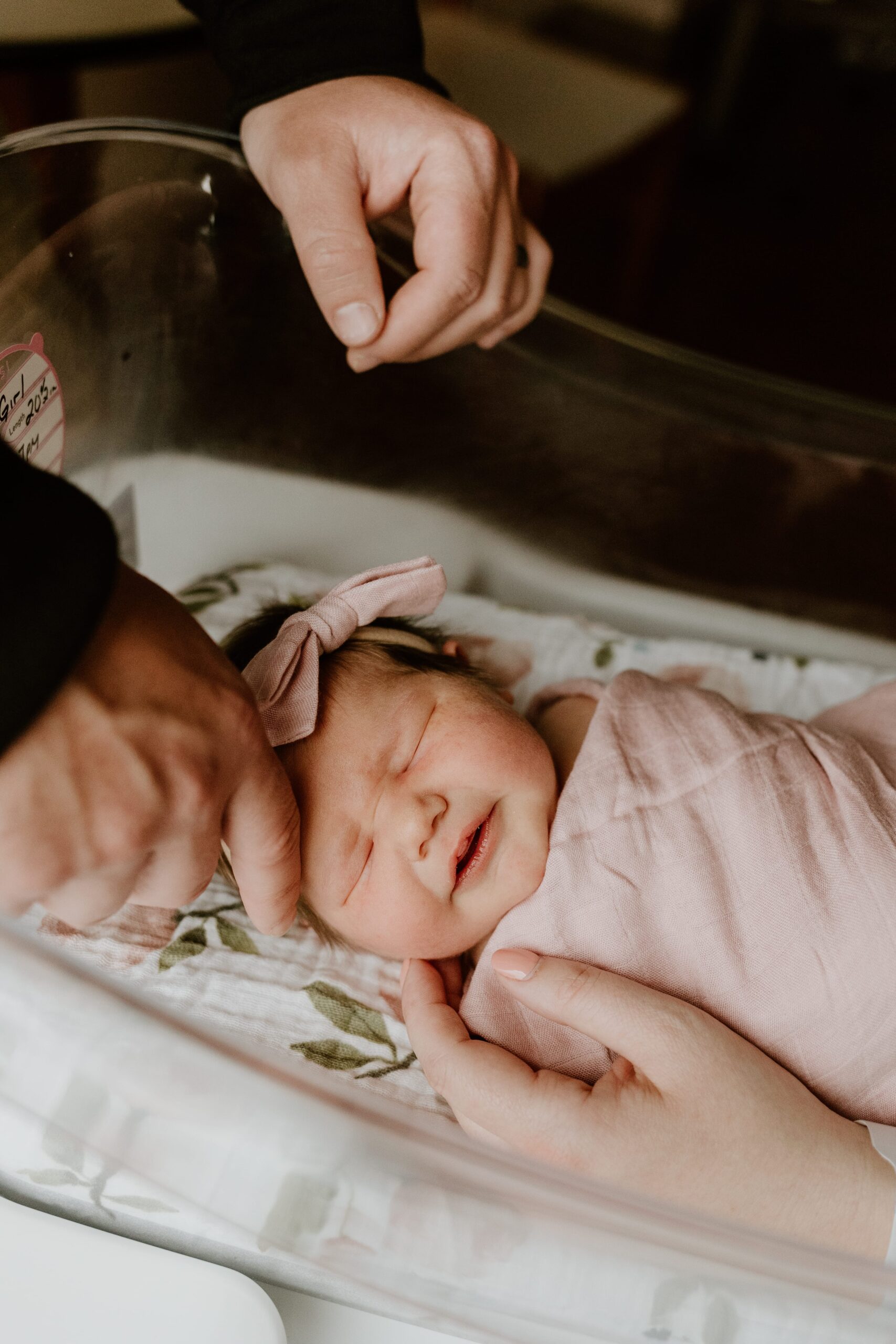 A newborn baby with a delicate pink bow headband is gently cradled in the hands of an adult at Vassar Brothers Medical Center. The baby is swaddled in a soft pink blanket and is peacefully asleep, showing a serene expression.