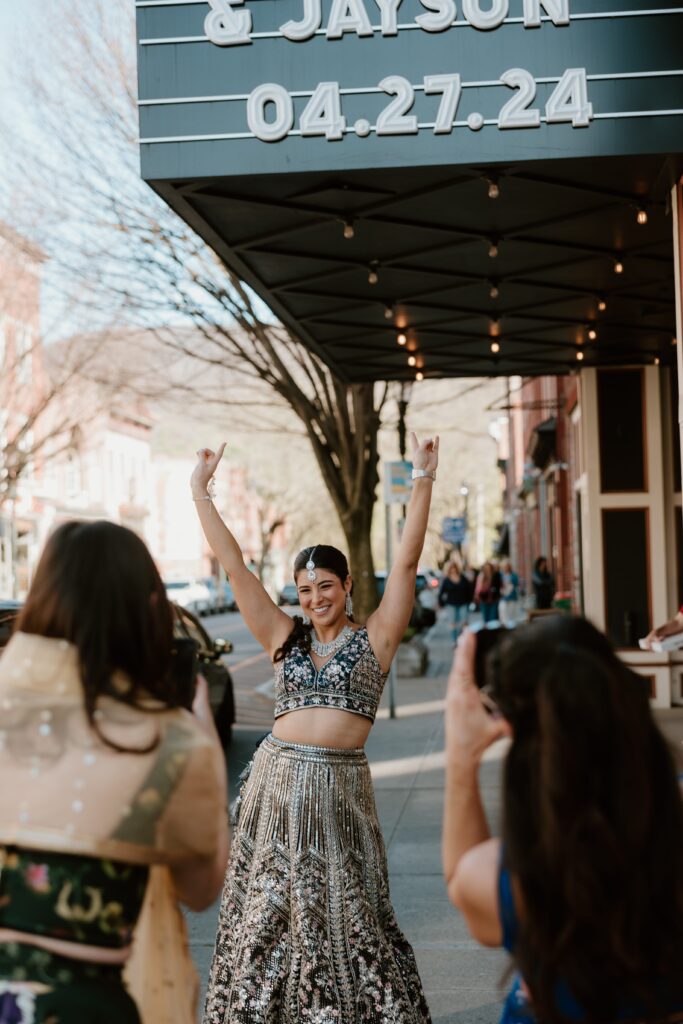 Hannah joyously raising her arms in celebration on a bustling street in Beacon, NY, under the marquee of Wonderbar announcing their wedding date. She is dressed in a beautifully detailed lehenga, smiling broadly as friends capture the moment, embodying the festive spirit of the occasion.