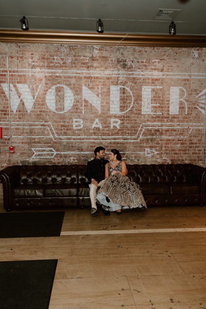 Hannah and Jayson sitting on a plush leather couch at Wonderbar, sharing a cozy moment against a rustic brick wall adorned with the 'Wonderbar' sign in bold white letters. Their affectionate gaze and smiles reflect a joyful and relaxed atmosphere during their celebration.