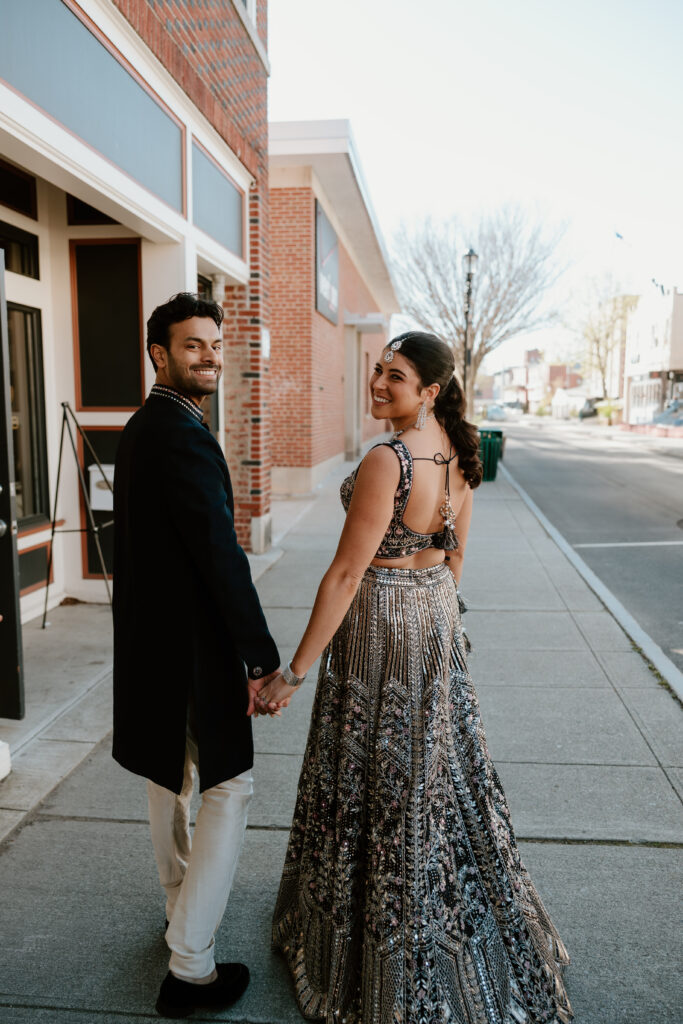 Hannah and Jayson walking hand in hand along a quiet street in Beacon, NY, outside Wonderbar. Hannah glances back, smiling, in her ornate lehenga, while Jayson looks forward, both radiating happiness. This candid moment captures the joy and anticipation of their wedding celebrations.