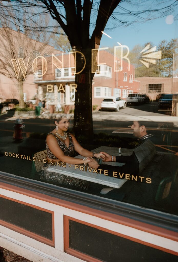 Hannah and Jayson seated at Wonderbar, seen through a window reflection that overlays text advertising cocktails, dining, and private events. The couple holds hands, sharing a private moment, framed by the warm interior and the bustling street outside.
