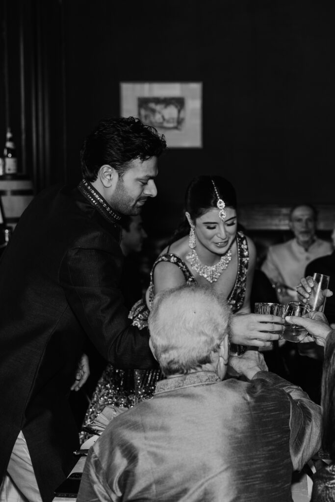 Jayson and Hannah joyfully engaging with guests during a toast at their dinner rehearsal, both dressed in traditional attire. Jayson in a dark embroidered sherwani and Hannah in a bejeweled lehenga, sharing a toast with an elderly guest in a relaxed, festive atmosphere.