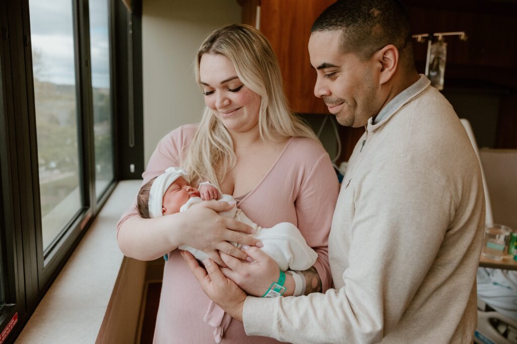 Mother and father lovingly holding their newborn baby by the window at Vassar Hospital.