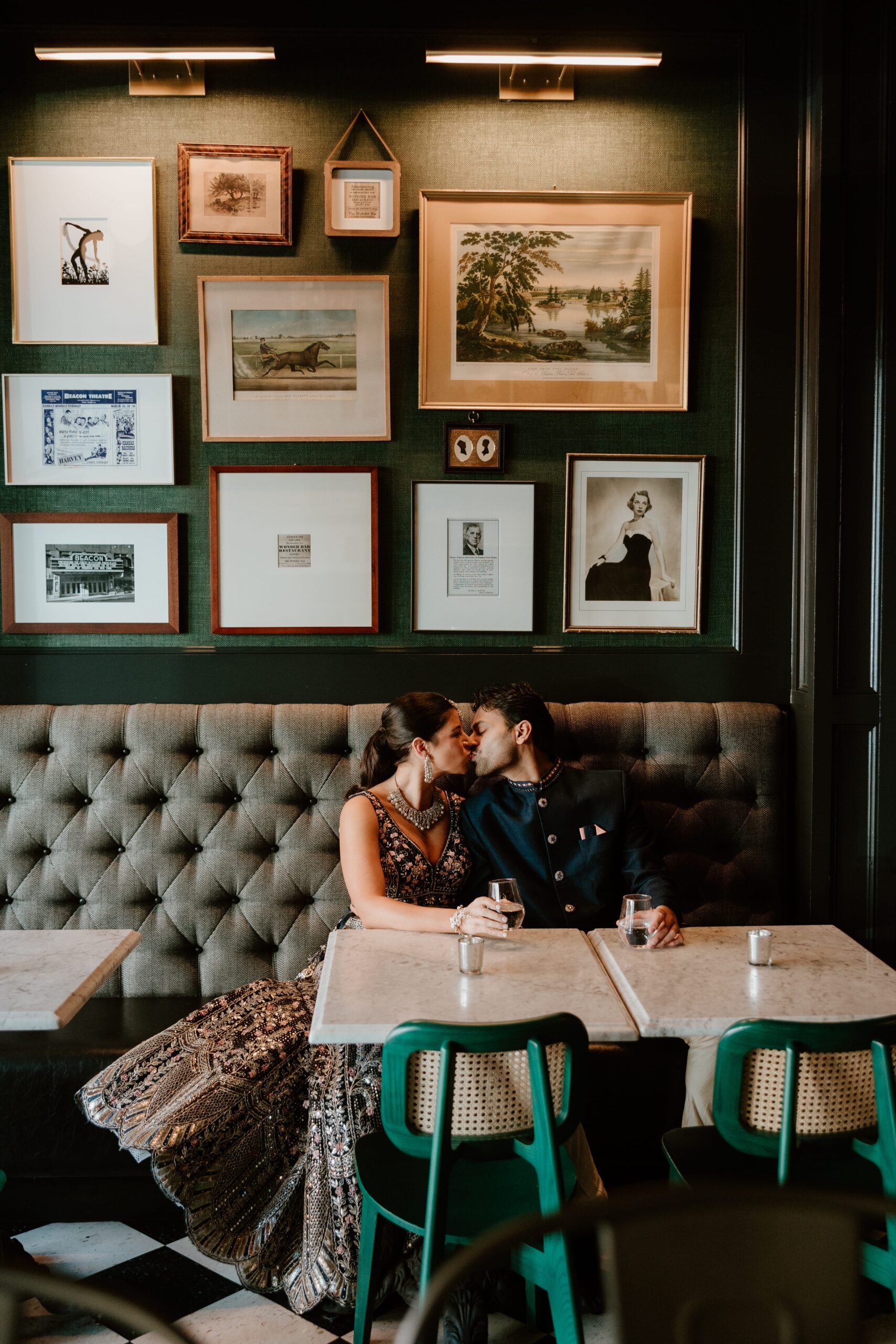 A couple sharing a romantic kiss at a table in Wonderbar, Beacon, with a backdrop of an elegant wall adorned with a variety of framed artworks and photographs, under warm lighting. They are seated on a tufted leather booth, surrounded by chic decor and vibrant green chairs, capturing the intimate ambiance of their dinner rehearsal.