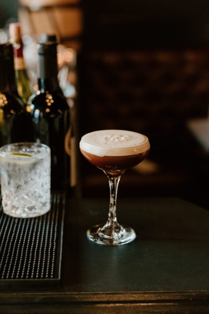 A sophisticated cocktail with a creamy froth topping and sprinkled garnish, presented on a dark bar counter alongside a glass of ice water. Behind it, a selection of wine bottles adds to the cozy, inviting atmosphere of the bar.