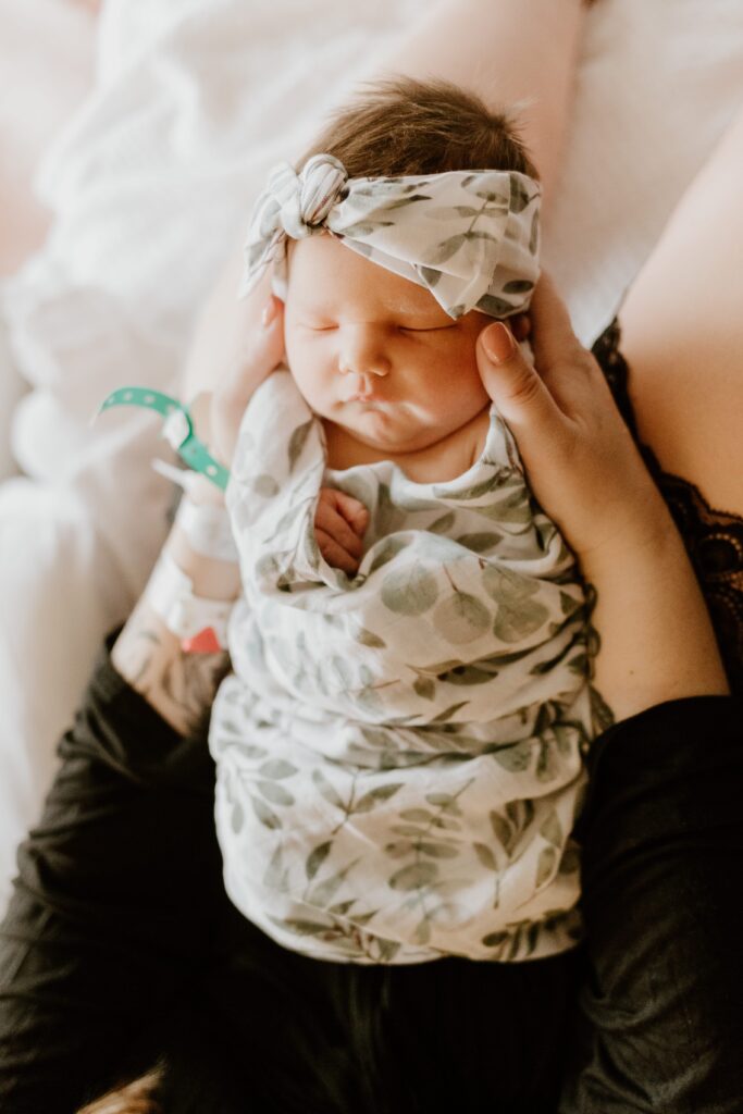 Newborn baby swaddled in a leaf-patterned blanket with a matching headband, resting in the arms of a person, in a setting of Vassar Hospital's labor and delivery room.