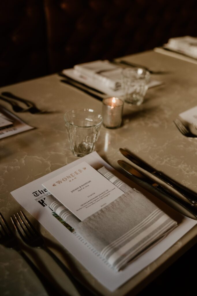 An elegantly set dining table at Wonderbar, Beacon, featuring a textured gray tablecloth, neatly arranged silverware, and a menu titled 'The H' with a detailed list of courses for Jayson and Hannah's rehearsal dinner, accented by soft candlelight and a serene atmosphere.