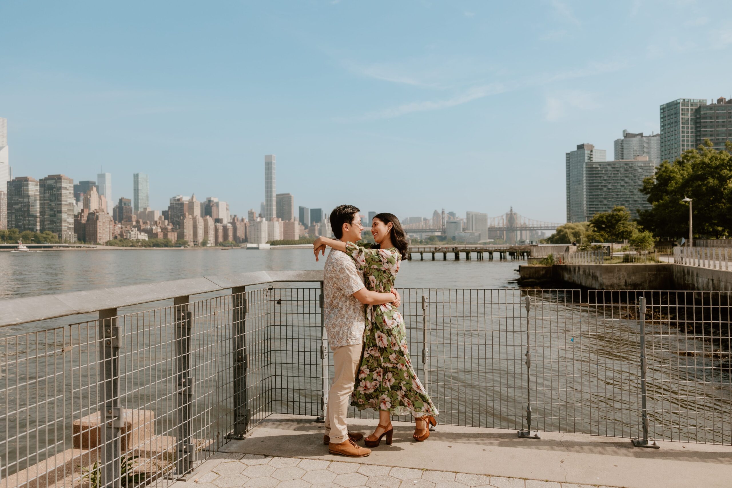 Sam and Mark embracing on a waterfront pier with the NYC skyline in the background at Gantry Plaza State Park during their engagement session.